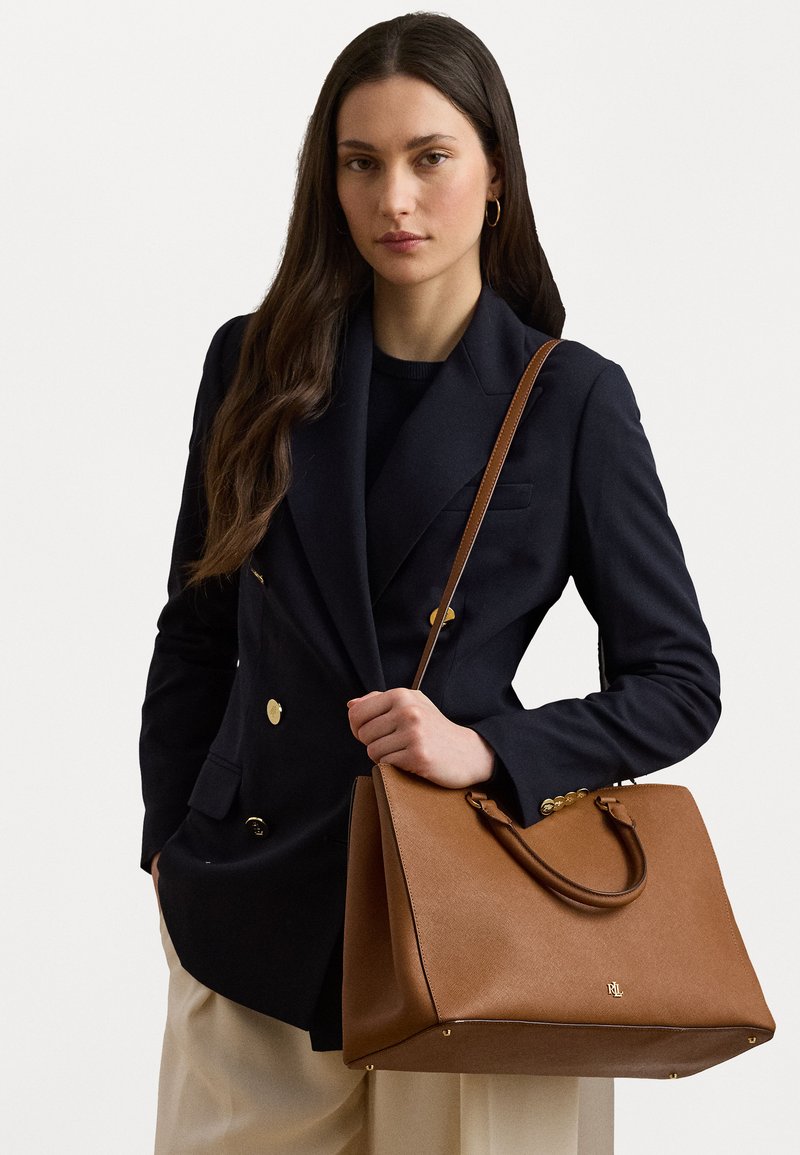 Porjavna usnjena torbica v tan barvi s strukturirano obliko, z zlatimi kovinskimi dodatki in dvema ročajema. Površina ima teksturiran zaključek in detajl z logotipom.