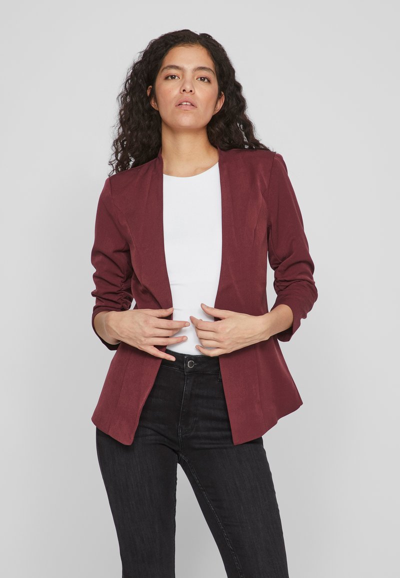 Bordowa marynarka o dopasowanym kroju, z usztywnionymi ramionami, bez klap. Posiada długie rękawy i gładką fakturę. Noszona na białej bluzce.