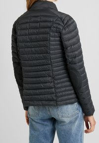 En person ses fra ryggen iført en sort, pufjakke med vandret syninger og albueforstærkninger, parret med lyseblå jeans.
