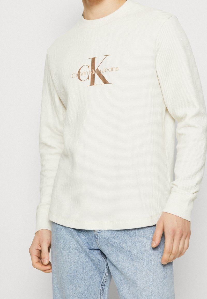 Personne portant une chemise crème à manches longues avec le logo "Calvin Klein Jeans" marron et un jean bleu clair, debout devant un fond uni.