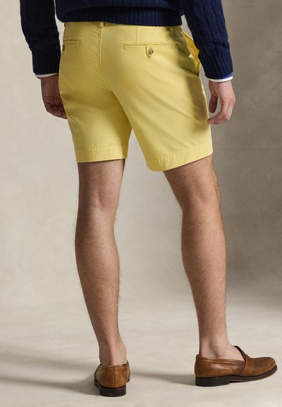 Žlté bavlnené šortky s vreckami, s hladkou textúrou a priliehavým strihom. Nosia sa s hnedými koženými mokasínami. Zobrazený zadný pohľad.