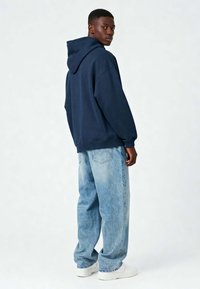 Jonge man in een marineblauwe hoodie, lichtblauwe baggy jeans en witte sneakers staand en over zijn schouder kijkend tegen een effen achtergrond.