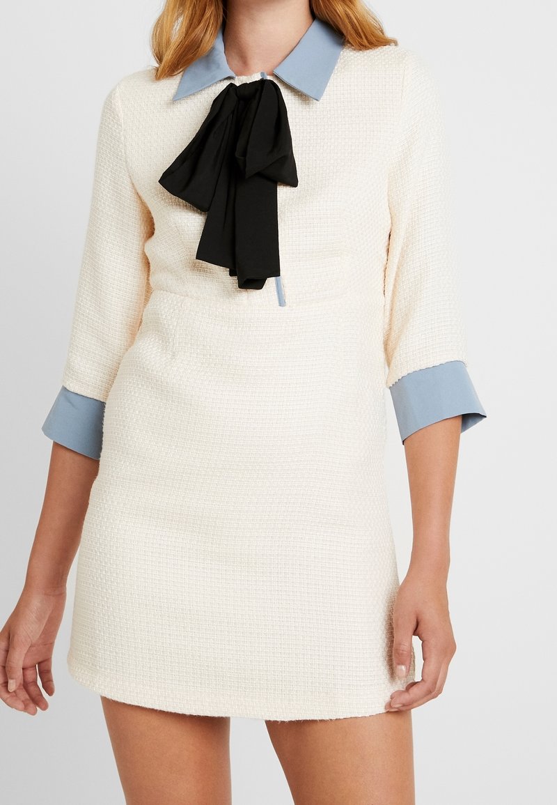 Femme portant une robe texturée crème avec un col et des poignets bleu clair, ornée d'un grand nœud noir noué au cou.