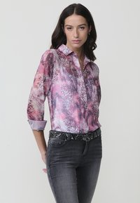 Camisa de manga larga con botones en estampado animal de color rosa y púrpura con un acabado brillante, combinada con pantalones vaqueros oscuros y un cinturón estampado.