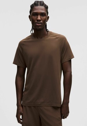 Mand med flettet hår iført en brun kortærmet skjorte og matchende brune bukser mod en ensartet lys baggrund.