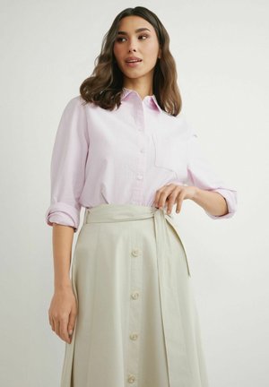 Kobieta w jasnoróżowej koszuli na guziki z podwiniętymi rękawami oraz w beżowej spódnicy z wysokim stanem, z guzikami i zawiązanym paskiem.