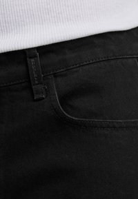 Detailný záber na vrecko čiernych džínsov a pútko na opasok pod bielym rebrovaným tričkom, ktorý zvýrazňuje textúru látky a detaily šitia.