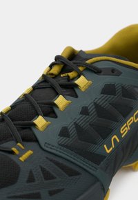 Σκούρο γκρι αθλητικό παπούτσι με κίτρινες λεπτομέρειες, υφή στην επιφάνεια, έντονο μοτίβο πέλματος και σχέδιο με κορδόνια. Διαθέτει επένδυση με μαξιλάρι στο πέλμα.