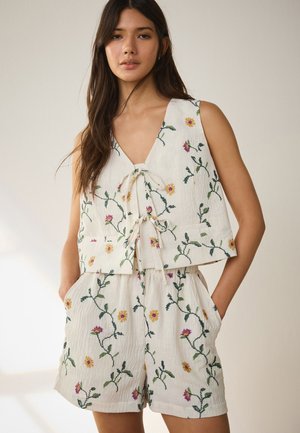 Femme portant un haut blanc sans manches et un short assorti avec une broderie florale verte et jaune, debout les mains dans les poches.