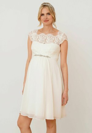 Donna con abito bianco in pizzo fino al ginocchio con maniche a mantella e fascia per capelli decorata con gioielli, in piedi davanti a uno sfondo chiaro e semplice.