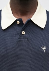 Laivastonsininen teksturoitu poolopaita, jossa on kermaisenvärinen kaulus, kaksi vaaleaa nappia ja sydämenmuotoinen kirjailu rintapielellä.