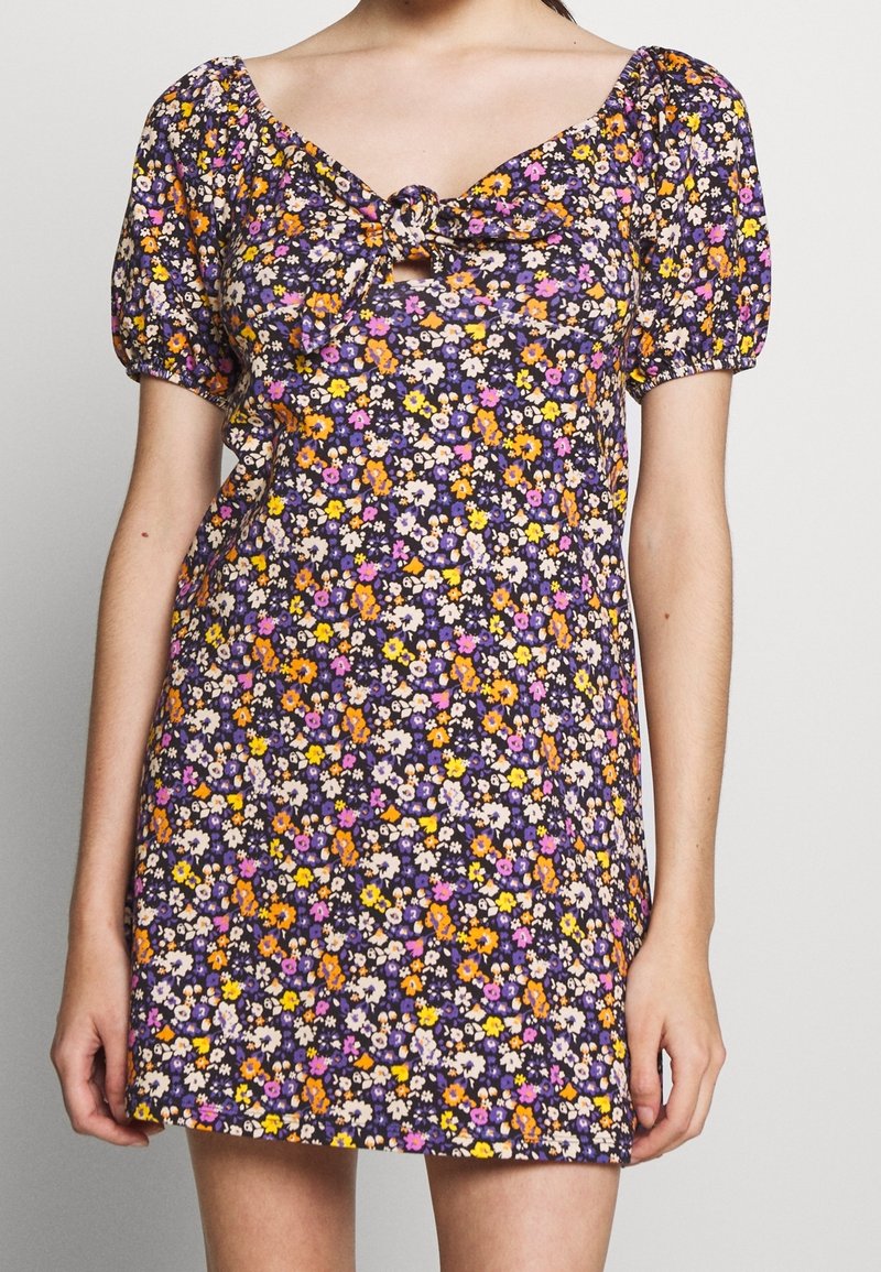 Kwiatowa mini sukienka na ciemnogranatowym tle, z fioletowymi, żółtymi i pomarańczowymi kwiatami. Krótkie, bufiaste rękawy oraz wiązany detal z przodu.