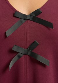 Camiseta burdeos con escote en V y tres lazos de cinta negra en la parte delantera. La tela es suave y con un ligero estiramiento.