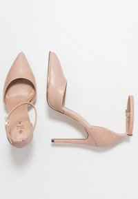 Nude, spitz zulaufende High Heels mit einem glatten Lederobermaterial und einem strukturierten Fersenriemen. Dünner Stiletto-Absatz und goldene Details.