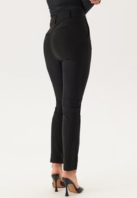 Zwarte op maat gemaakte broek met een gladde textuur, slim fit en tailleband. Draagt met zwarte hoge hakken. Strak, minimalistisch ontwerp.