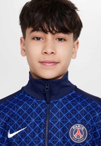 Veste de sport bleu marine avec un motif géométrique, col montant, fermeture éclair complète et un logo PSG rond sur la poitrine gauche. Fabriquée en tissu lisse.