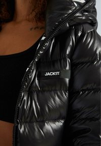 Černá péřová bunda s lesklými částmi, vybavená zipovým zapínáním a malou zelenou nálepkou s nápisem "JACK1T" na horní části rukávu.