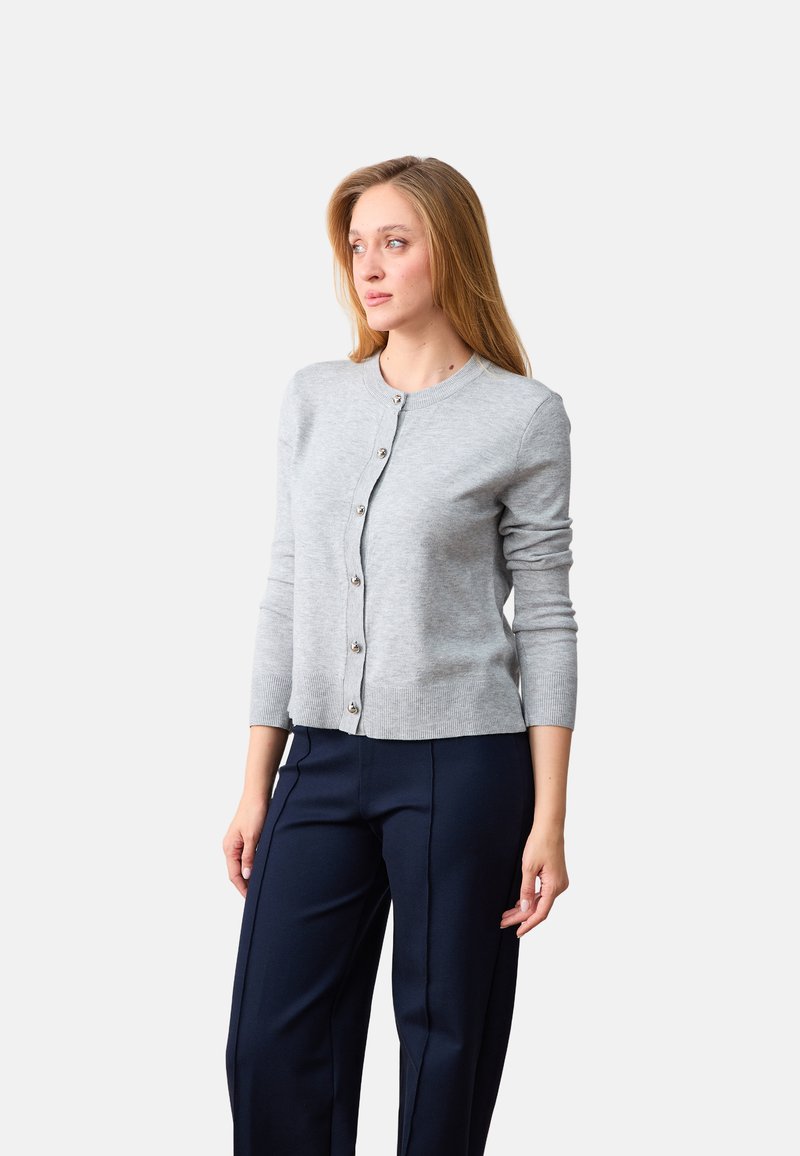 Femme aux longs cheveux blonds portant un cardigan gris clair boutonné et un pantalon bleu marine foncé, regardant vers la gauche sur un fond uni.