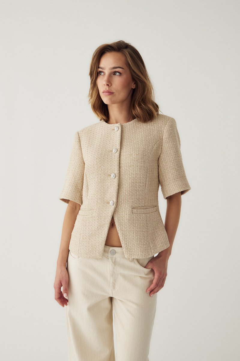 Femme portant une veste beige texturée à manches courtes boutonnée et un pantalon beige clair, debout avec une main dans la poche, regardant sur le côté.