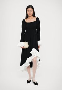 Vestido negro con mangas largas, corsé fruncido y acentos de volantes blancos en el dobladillo y las mangas. Modelo sosteniendo un pequeño bolso de color crema.