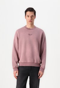 Hellrosa Sweatshirt mit gerippten Bündchen und Saum, ausgestattet mit einem Rundhalsausschnitt und besticktem Text. Kombiniert mit dunklen Jeans. Weicher Stoff. 