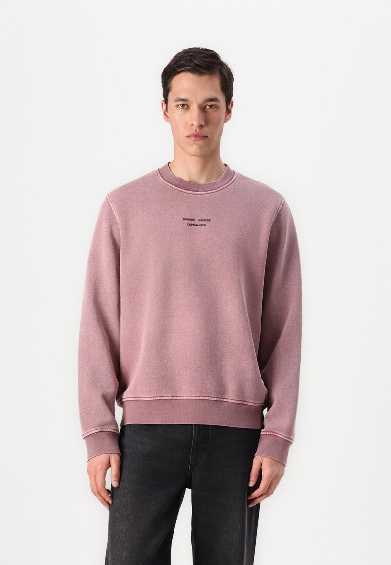 Hellrosa Sweatshirt mit gerippten Bündchen und Saum, ausgestattet mit einem Rundhalsausschnitt und besticktem Text. Kombiniert mit dunklen Jeans. Weicher Stoff. 