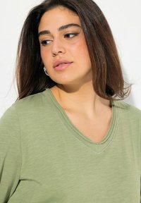 Světle zelené tričko s véčkovým výstřihem, jemnou texturou a decentním stříbrným prošíváním podél límce; vystavené na jednoduchém bílém pozadí.