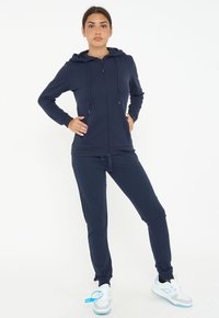 Felpa navy con zip e cordini, abbinata a pantaloni jogger, realizzati in tessuto morbido. Presenta una vestibilità rilassata e polsini a coste, abbinata a scarpe da ginnastica.