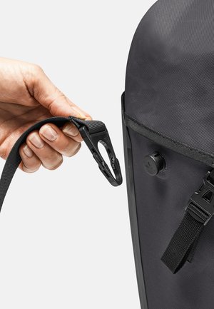 Zaino nero con un tessuto testurizzato. Una mano tiene una cinghia con un clip di plastica, mostrando il suo design e i dettagli di attacco.