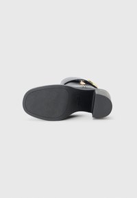 Zwarte leren sandaal met een platte zool, voorzien van gouden hardware accenten en een gestructureerde zool. De hak is gematigd en iets taps toelopend.