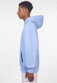 Helle blaue Kapuzenjacke aus weichem Stoff, mit großer Kapuze, Fronttasche und gerippten Bündchen. Textdesign auf der Vorderseite. Lässiger, lockerer Schnitt.