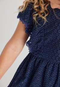 Granatowa sukienka z fakturowanej tkaniny z ażurowym wzorem, z falbaniastymi rękawami i zgromadzonym w pasie krojem.
