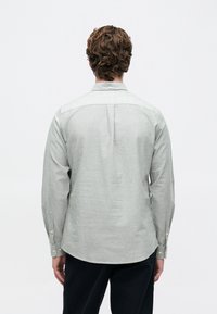 Homem com cabelo cabelo cacheado, vestindo uma camisa de botão de mangas longas cinza claro e calças pretas, visto de costas contra um fundo branco liso.