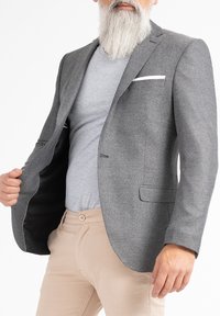 Blazer grigio con texture, colletto a lancia, taschino singolo con accentuazione bianca e foderato di nero. Indossato sopra una camicia grigia e pantaloni beige.