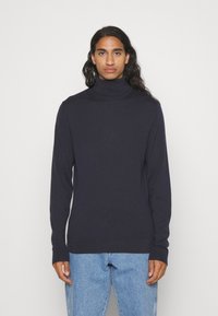 Maglione a collo alto di colore navy in un morbido tessuto a maglia, con maniche lunghe e una vestibilità affusolata, abbinato a jeans azzurri chiari.