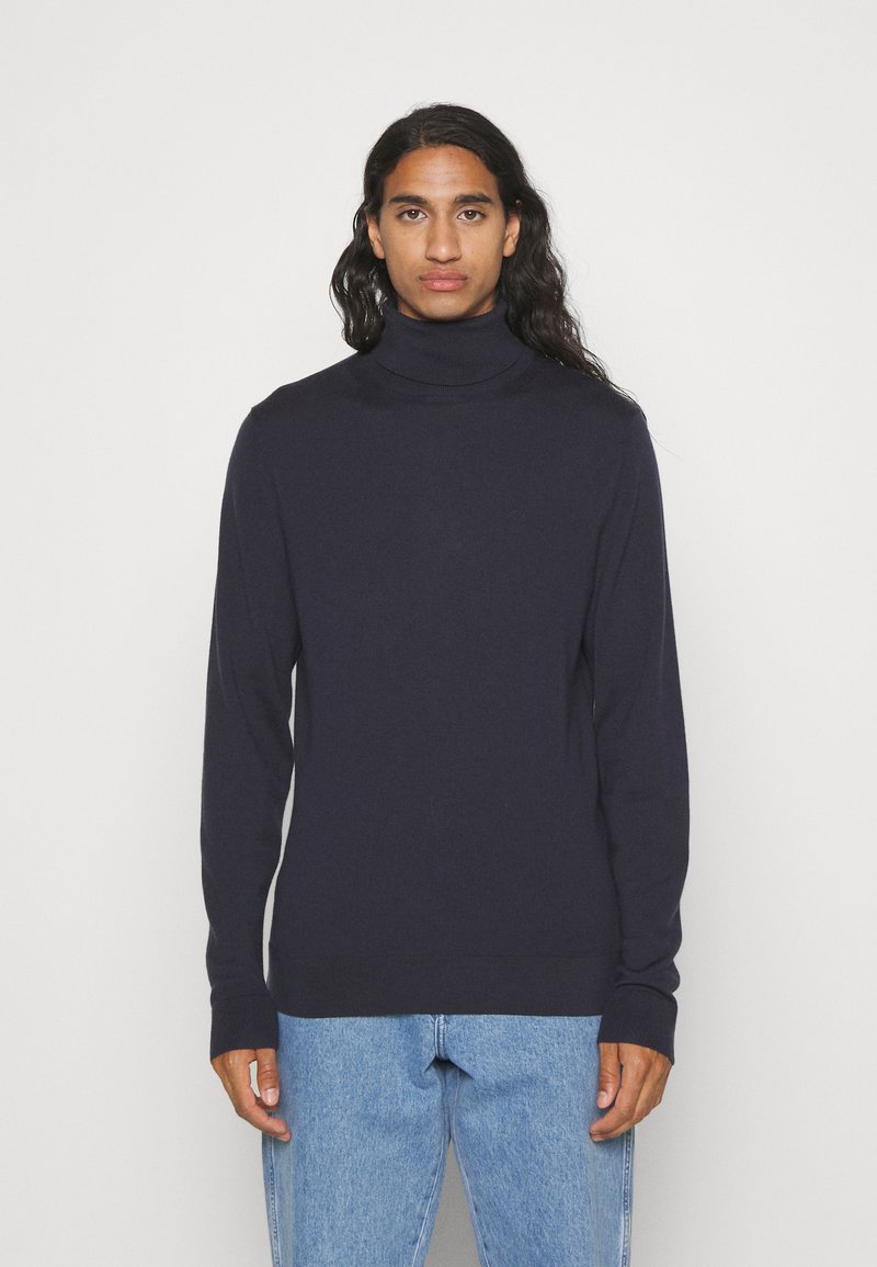 Maglione a collo alto di colore navy in un morbido tessuto a maglia, con maniche lunghe e una vestibilità affusolata, abbinato a jeans azzurri chiari.
