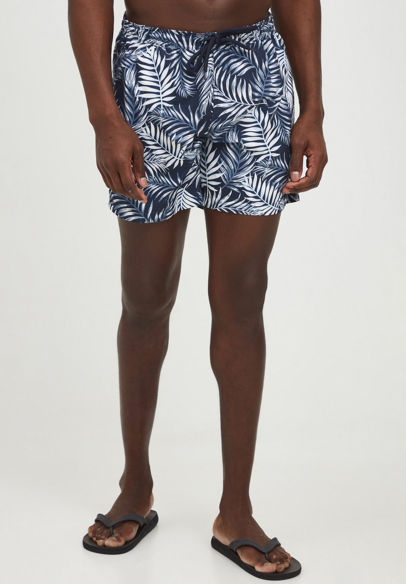 Marinblå badshorts med vitt mönster av palmblad, elastisk midja med dragsko och lättviktigt tyg. Bärs med svarta flip-flops.
