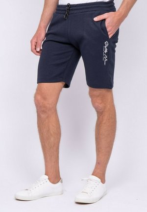 Homme portant un short en coton bleu marine avec des poches latérales, un texte blanc sur la cuisse gauche et des baskets blanches sur fond blanc.