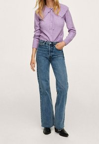 Donna che indossa un maglione lavorato a maglia lavanda con un ampio colletto, jeans blu a vita alta e stivali neri fino alla caviglia, in piedi con una mano in tasca.