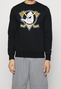 Svart sweatshirt med en stor tecknad spöke och korsade hockeyklubbor i vitt, guld och gult på framsidan. Tillverkad av bomullsmaterial.