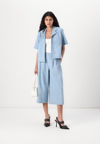 Completo a due pezzi in tessuto blu chiaro con strisce bianche verticali. Camicia corta a maniche corte e con taschino, pantaloni larghi plissettati e scarpe con tacco alto.