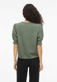 Vrouw met krullend haar, gekleed in een olijfgroene korte-mouw gebreide top met opgepofte mouwen en zwarte broek, met haar rug naar de camera toe.