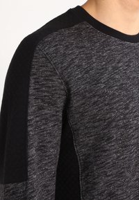 Pull en tricot gris avec épaules texturées noires. Dispose d'un col rond et d'une finition en tissu lisse avec des variations subtiles de couleur.