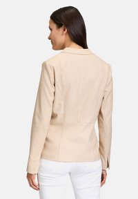 Beżowa marynarka o dopasowanym kroju, z klapami, szwami na ramionach i guzikami przy mankietach. Miękka faktura, zestawiona z białymi spodniami.