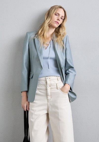 Jasnoniebieska marynarka o dopasowanym kroju i błyszczącej teksturze, zestawiona z żebrowanym topem. Beżowe spodnie z wysokim stanem z widocznymi guzikami i kieszeniami.