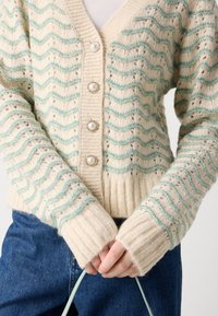 Cardigã de malha em creme com padrões ondulados em teal. Apresenta punhos canelados e quatro fechos decorativos com botões de pérola. Tecido suave e texturizado.