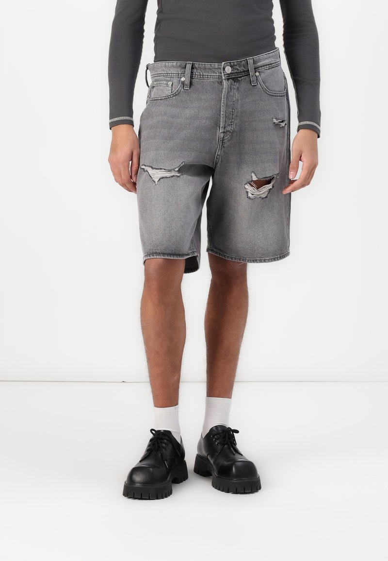 Shorts en denim gris avec des détails effilochés, des poches avant, une fermeture à bouton et une coupe décontractée, assortis à des chaussures noires à semelle épaisse.