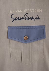 Beiger Textilstoff mit einer blauen Tasche, die einen Knopf hat. Das Design umfasst bedruckten Text: "JAN VANDERS TORM" und "Sean Scandinavia."