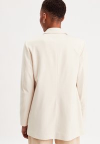 Lys beige blazer med en struktureret design, der har en enkelt slids, lange ærmer og subtile syninger. Glat stoftekstur.