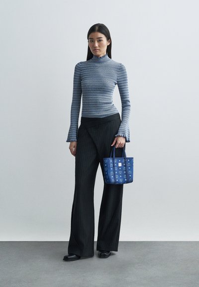 Γυναίκα με ριγέ μπλε μπλούζα μακρυμάνικη, μαύρο παντελόνι φαρδύ και μαύρα παπούτσια, κρατώντας γαλάζια τσάντα με μοτίβο σε απλό φόντο.